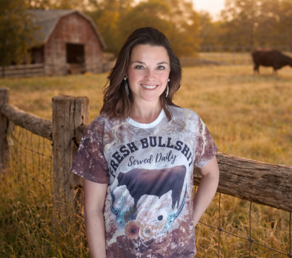 Woman wearing a graphic t-shirt in a rural setting with a barn and cow in the background.